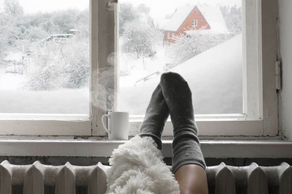 Cozy winter still life: woman legs in warm woolen socks under shaggy blanket and mug of hot beverage on old windowsill against snow landscape from outside.