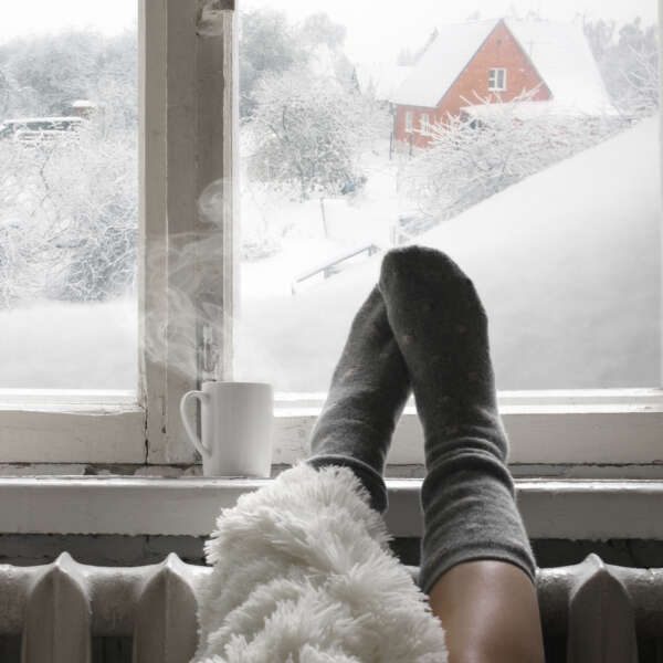 Cozy winter still life: woman legs in warm woolen socks under shaggy blanket and mug of hot beverage on old windowsill against snow landscape from outside.